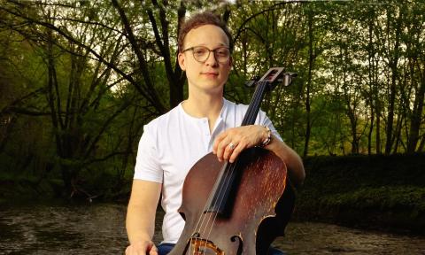Cellist Ben Solee sitting in a wooded area with his cello