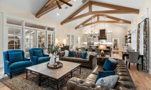 This great room, as seen from the television wall, has a large seating area, dining table, and white kitchen, enhanced with open wood beams