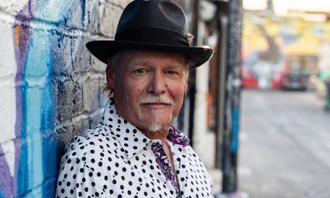 Jimmy Carpenter wears a black hat and leans on a colorful brick wall. 