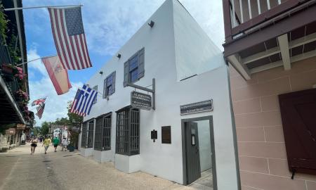The outside of the Avero House / St. Photios Shrine from St. George Street on a summer day. Tourists walk below Greek and American flags