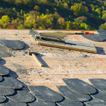 A full color image of a roofer's tools (small wooden hammer and tiles and scraper) on a semi-tiled roof. The shingles (tiles) are ovular and gray. Trees can be seen past the roof ridge in the background.
