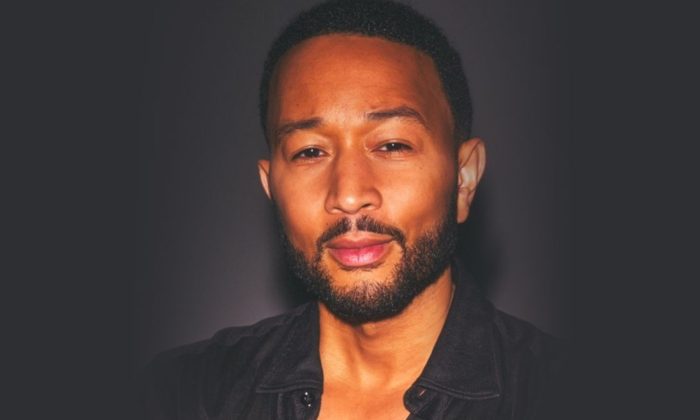 John Legend wears a black shirt and poses in front of a black backdrop.